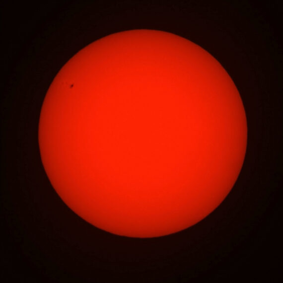 Disco solare rosso su fondo nero con una piccola macchia solare a sinistra e dati di scatto in basso.